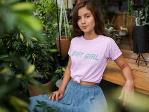 brown haired woman wearing pink t shirt with soft girl text and jean dress skirt sitting on bench surrounded by green foliage and potted plants